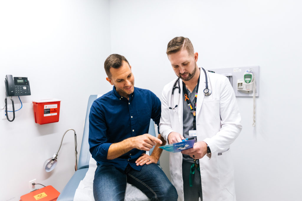 A man wearing a blue shirt learns about HIV treatment and primary care at Metro Inclusive Health. A HIV primary care provider holds information about PrEP and medical care.