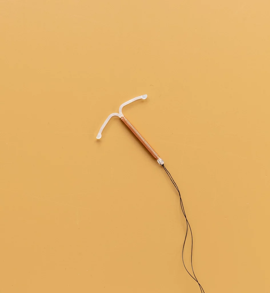 A single, copper intrauterine device (IUD) against a solid mustard-yellow background. The T-shaped plastic frame of the IUD and its fine copper coil windings are clearly visible, along with the black removal threads extending from the base. The image captures the IUD in a centralized position, providing a clear and informative view of this contraceptive method.
