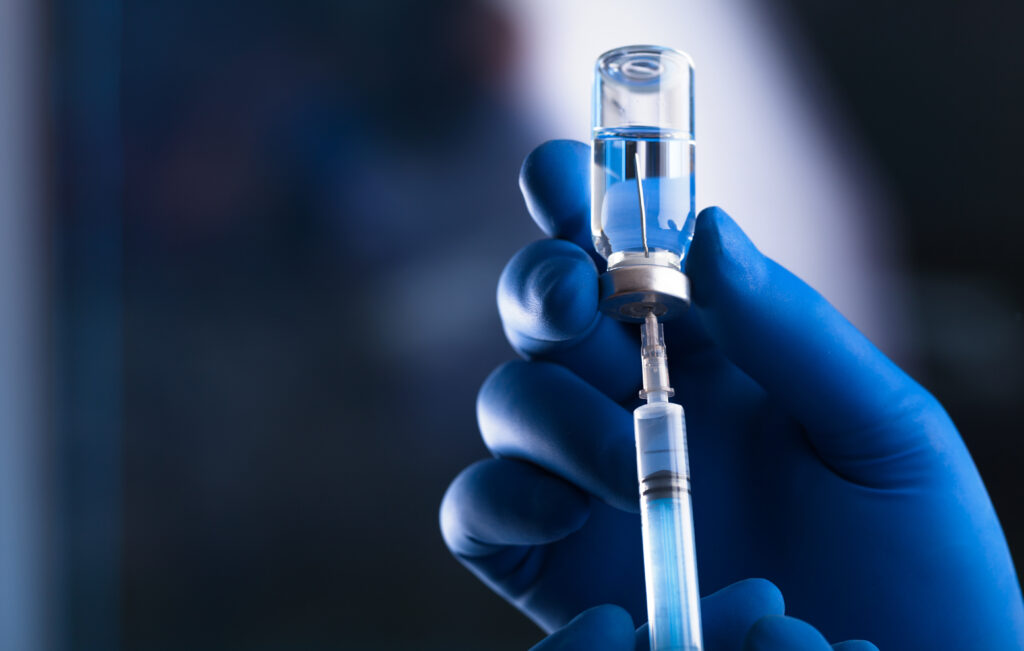 Close-up of a healthcare professional's hand in blue surgical gloves, holding a medical syringe with a vial. The syringe is being filled with a clear liquid from the vial, against a blurred background, emphasizing the precision and care in vaccine preparation.