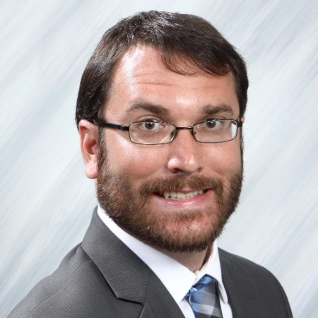 A professional portrait Brian Harris with dark hair and a beard, wearing glasses, a dark suit, and a blue-striped tie. The background is a gradient from white to light gray, suggesting a corporate setting.