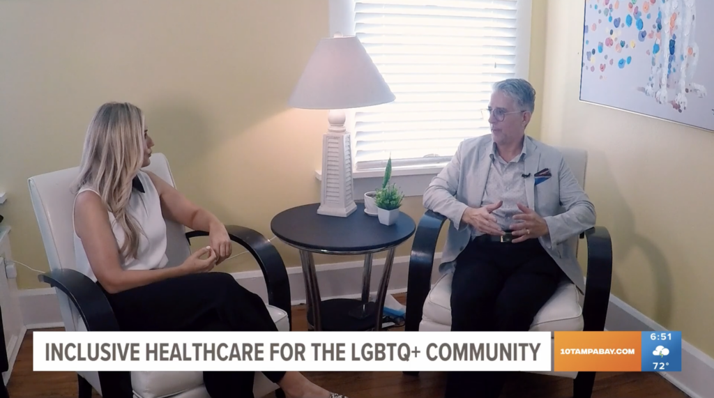 Screenshot from an interview, featuring a woman and a man seated across from each other in a warmly lit room with neutral tones. The banner at the bottom reads 'INCLUSIVE HEALTHCARE FOR THE LGBTQ+ COMMUNITY'. They appear to be in a discussion, which is being broadcasted on '10TAMPABAY.COM' with a weather update indicating '72°F' in the corner.