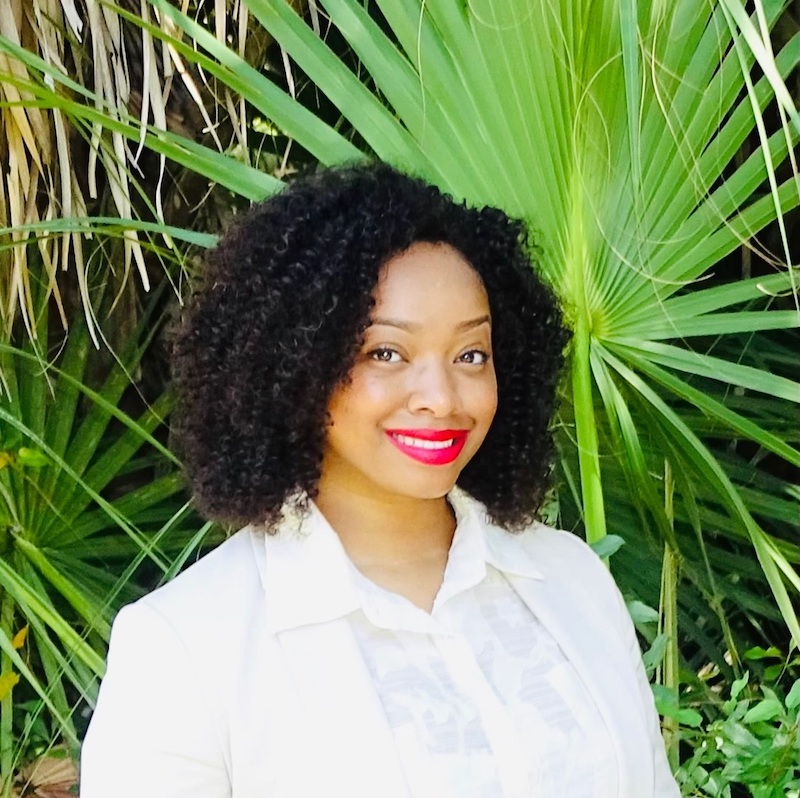 Headshot of Shericka Cunningham with curly hair, wearing a white blouse and bright red lipstick. She is posing in front of lush greenery, which adds a fresh and vibrant backdrop to the image.