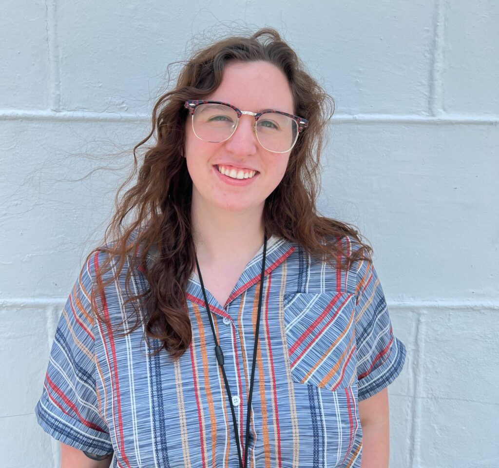 A smiling Laura Harvester with shoulder-length curly hair wearing clear-framed glasses and a striped shirt standing in front of a light-colored wall.