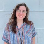 A smiling Laura Harvester with shoulder-length curly hair wearing clear-framed glasses and a striped shirt standing in front of a light-colored wall.