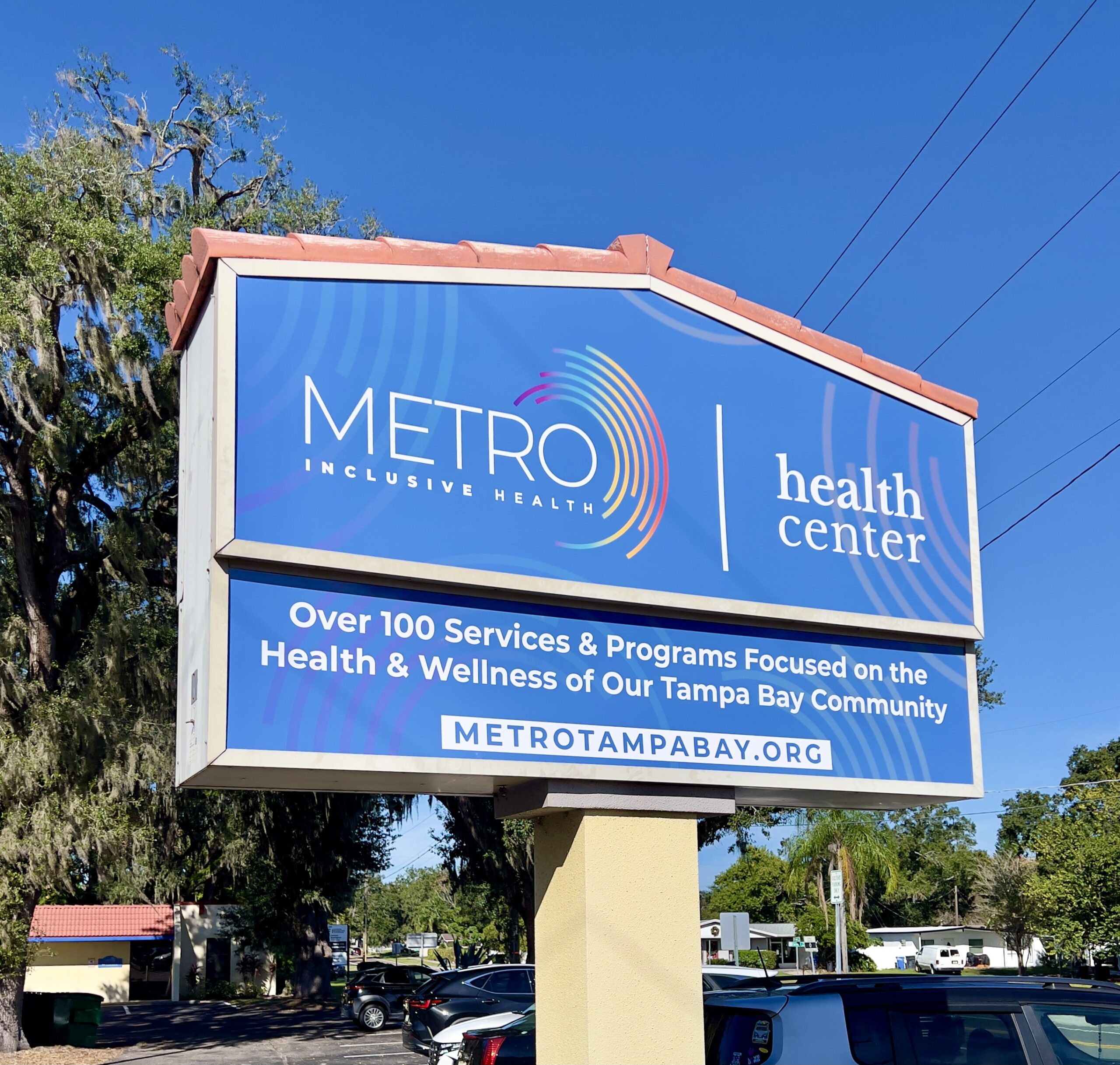 A roadside billboard for METRO Inclusive Health with a clear blue sky in the background. The sign features the METRO logo and reads 'health center' along with the message 'Over 100 Services & Programs Focused on the Health & Wellness of Our Tampa Bay Community' and the website 'METROTAMPABAY.ORG'.