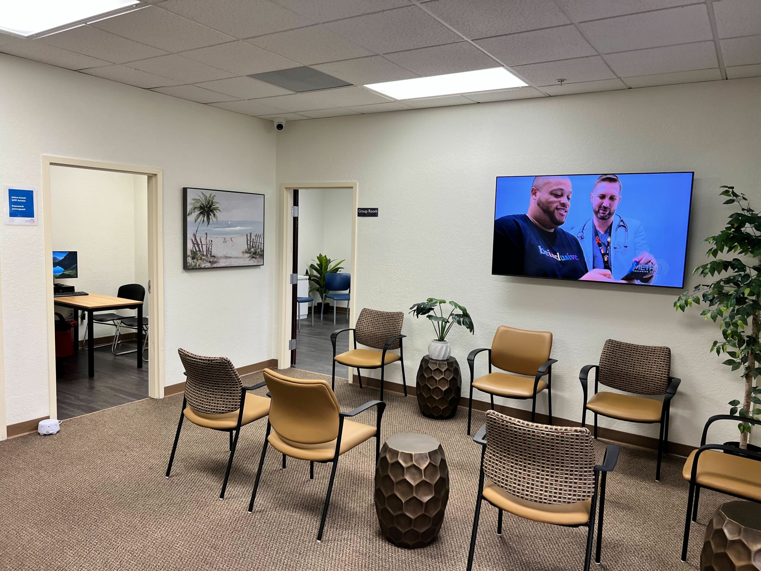A view of a modern and inviting clinic waiting room with comfortable chairs arranged neatly, a large TV screen showing healthcare content, tropical-themed wall art, and potted plants adding a touch of greenery. The room has a calm and professional atmosphere.