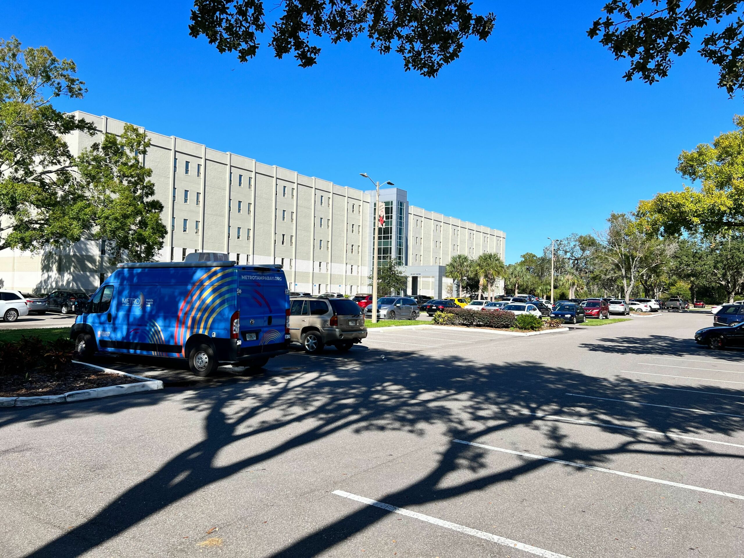 A METRO Inclusive Health branded van parked in a lot outside a multi-story medical facility on a sunny day. The van is wrapped with a colorful design featuring the METRO logo. Trees cast shadows over the parking area, creating a peaceful, suburban atmosphere.