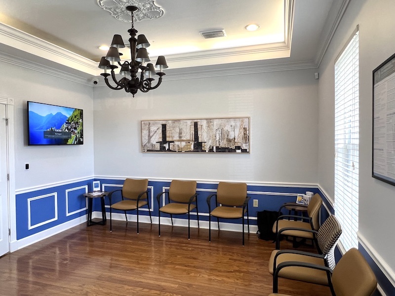 An elegant waiting room with hardwood floors, white walls with blue wainscoting, and a classic chandelier. The room is furnished with a row of taupe chairs along the wall, a decorative painting, and a mounted television displaying a nature scene.