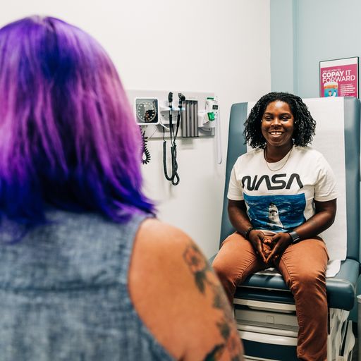A patient with a joyful expression is seated in a medical consultation room, facing a healthcare provider with purple hair, indicating a comfortable and inclusive healthcare environment at Metro Inclusive Health.