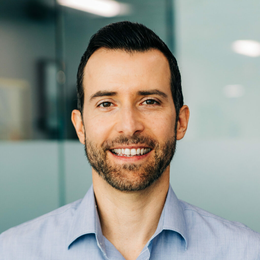 A headshot of Dr. Christopher Garnett with dark hair, a neatly trimmed beard, and a warm smile, wearing a light blue button-up shirt. The office setting in the background is blurred, focusing the viewer’s attention on the person’s friendly expression.