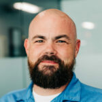 A portrait of Alex Monie with a full beard, wearing a blue denim shirt. His friendly gaze and confident posture convey a sense of approachability and professionalism.