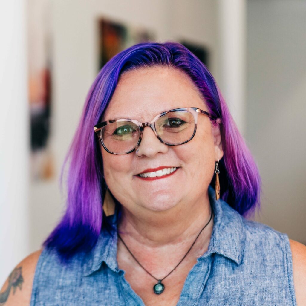 A close-up portrait of Lea Ann Kinney with vibrant purple hair and stylish round glasses, wearing a sleeveless denim top and a cheerful smile. They have a visible tattoo on their shoulder and are wearing drop earrings, projecting a creative and welcoming image.