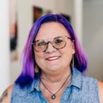 A close-up portrait of Lea Ann Kinney with vibrant purple hair and stylish round glasses, wearing a sleeveless denim top and a cheerful smile. They have a visible tattoo on their shoulder and are wearing drop earrings, projecting a creative and welcoming image.