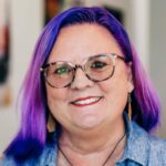 A close-up portrait of Lea Ann Kinney with vibrant purple hair and stylish round glasses, wearing a sleeveless denim top and a cheerful smile. They have a visible tattoo on their shoulder and are wearing drop earrings, projecting a creative and welcoming image.