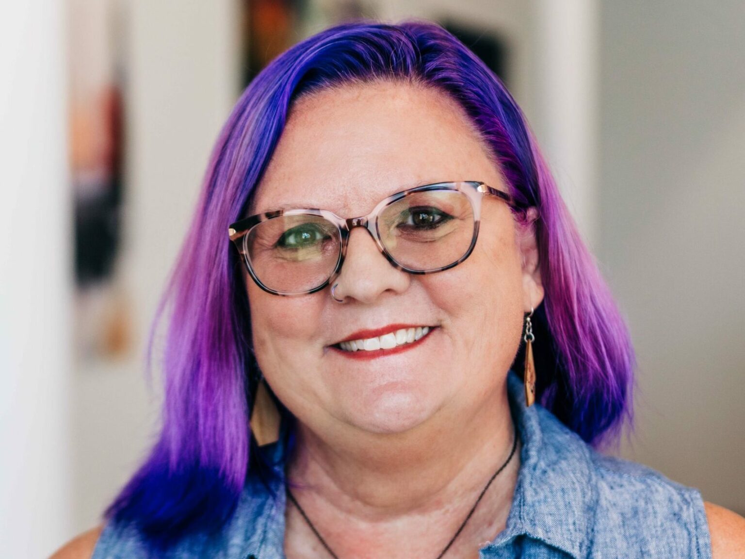 A close-up portrait of Lea Ann Kinney with vibrant purple hair and stylish round glasses, wearing a sleeveless denim top and a cheerful smile. They have a visible tattoo on their shoulder and are wearing drop earrings, projecting a creative and welcoming image.