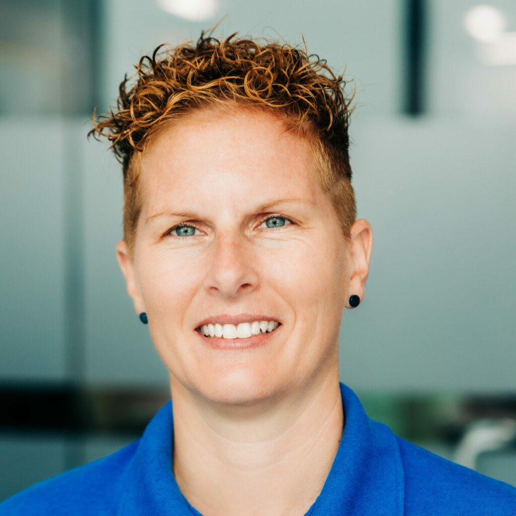 A close-up portrait of a smiling person with short curly hair highlighted with blonde tips, blue eyes, and a blue shirt. They have a confident, friendly demeanor and are wearing small black stud earrings, standing against a blurred indoor background.