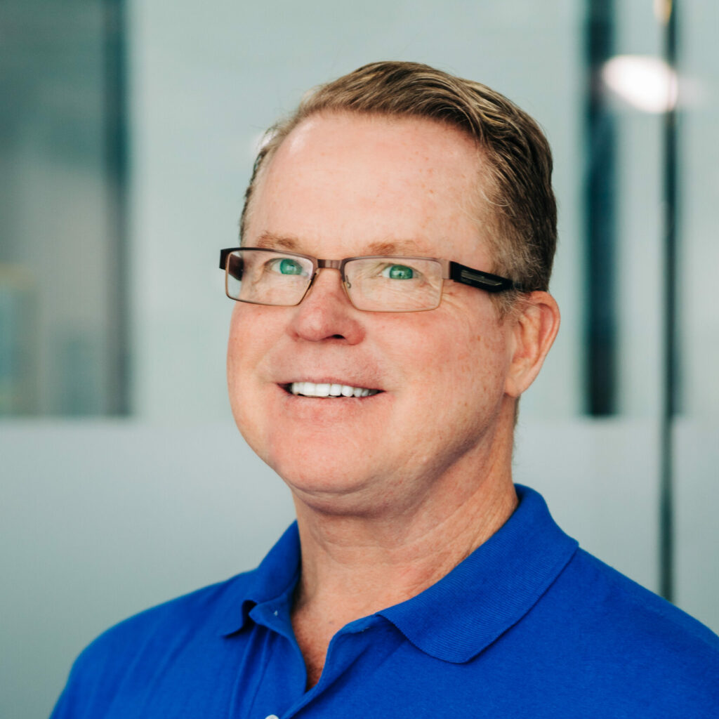 Portrait of Dr. Clyde Moreland with short, neat hair and glasses, wearing a blue polo shirt, smiling confidently in an office environment with a reflective glass surface in the background.