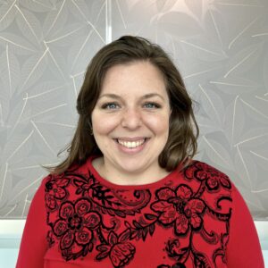 Headshot of Stephanie Whiteley shoulder-length brown hair and blue eyes, wearing a red sweater with detailed black floral embroidery. She is standing in front of a decorative wall with a leaf pattern, smiling warmly at the camera.