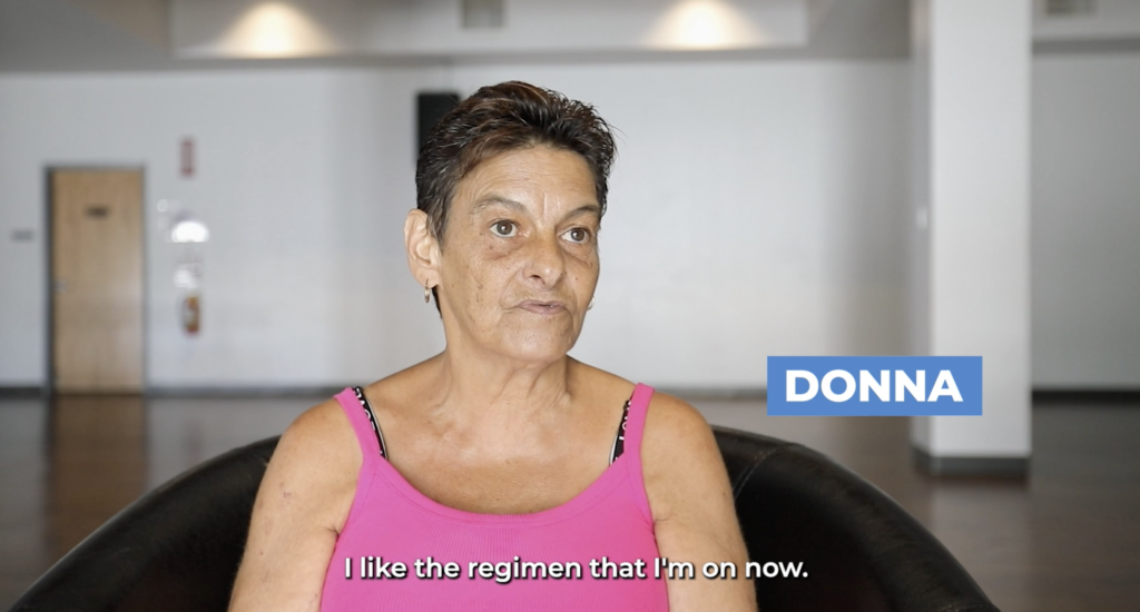 A woman named Donna is seated, looking directly at the camera, with a caption that reads 'I like the regimen that I'm on now.' She has short hair, is wearing a pink tank top, and there is a blurred interior background with a door and a light fixture.