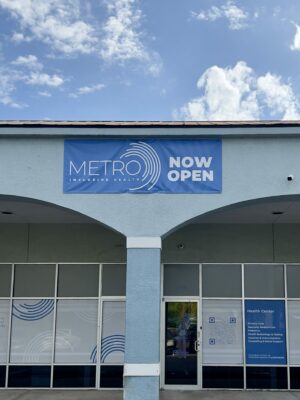 Exterior of our South St. Pete location. Blue walls and glass doors with white frosting and blue rings. Blue sign reading "METRO INCLUSIVE HEALTH NOW OPEN".