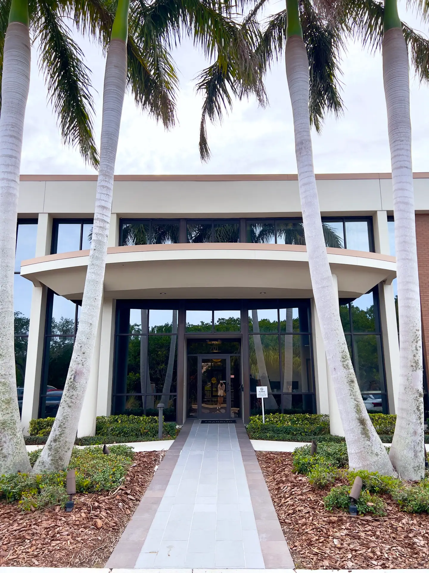 Exterior photo of the administrative office. Two royal palm trees on both sides of the walkway leading up to the glass front doors. Orange trim and tan walls.