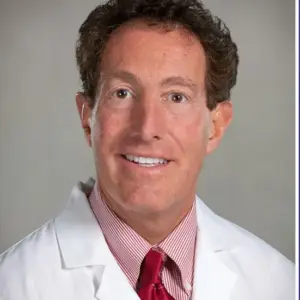 Headshot of Dr. Michael Nieder with curly brown hair, wearing a white lab coat, a red and white striped shirt, and a red tie. The background is a neutral gray, highlighting the professional demeanor of the subject.
