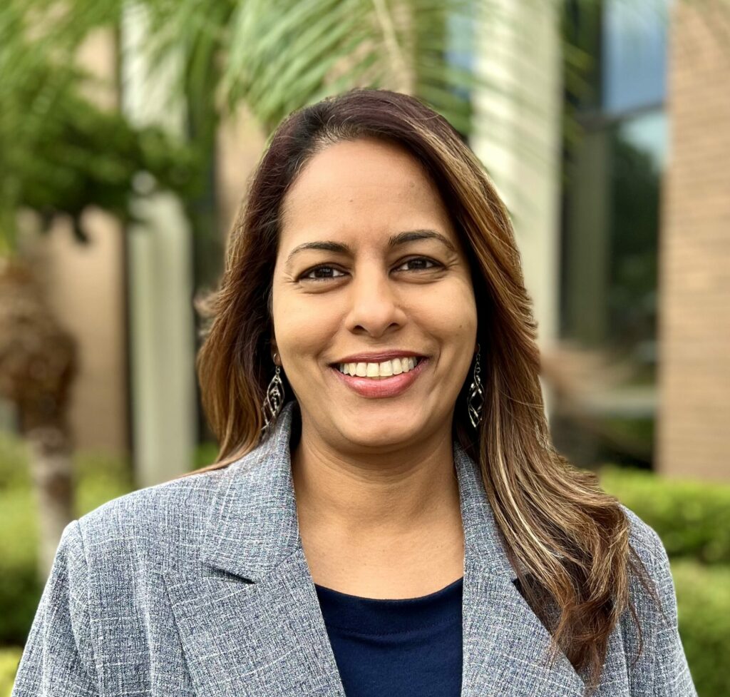 A professional headshot of Priya Rajkumar with long brown hair, wearing a grey blazer over a navy blue top and silver earrings. The background is softly blurred with greenery and part of a building, suggesting an outdoor setting.