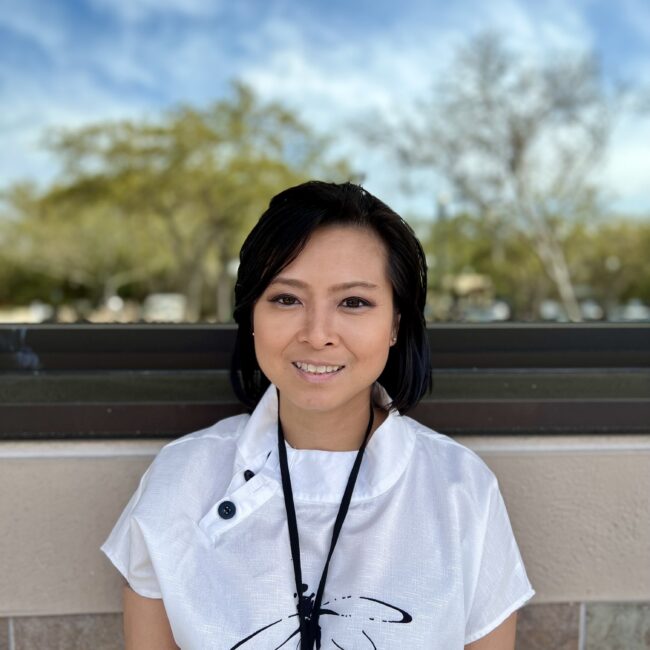 Portrait of Arie Ho with shoulder-length black hair, wearing a white shirt with a black tie detail at the neckline. She wears a lanyard around her neck and is pictured outdoors with trees and a blue sky with light clouds in the background.