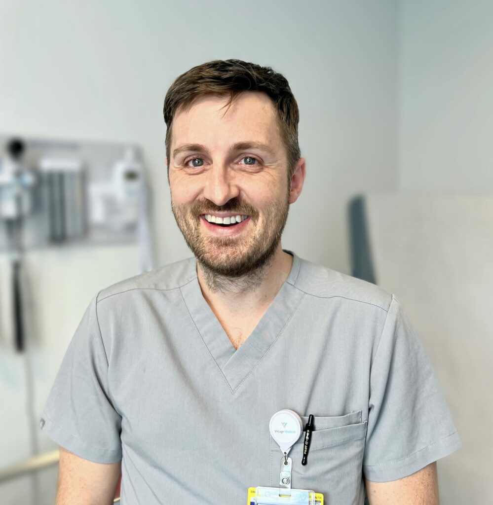 A smiling male healthcare professional wearing a grey medical scrub top and an employee ID badge from METRO Inclusive Health. He appears cheerful and approachable, standing in a clinical setting with a soft blue background that suggests a calm and welcoming environment. His badge and lanyard include a rainbow design, indicating a commitment to diversity and inclusivity.