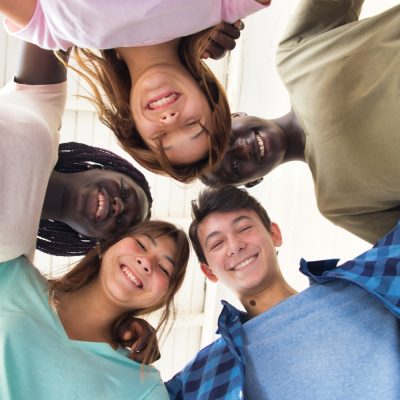 A heartwarming view from below as a diverse group of smiling young people forms a circle, their heads close together, looking down towards the camera. The image captures a moment of friendship and unity under a bright, clear sky, conveying a sense of inclusivity and community.