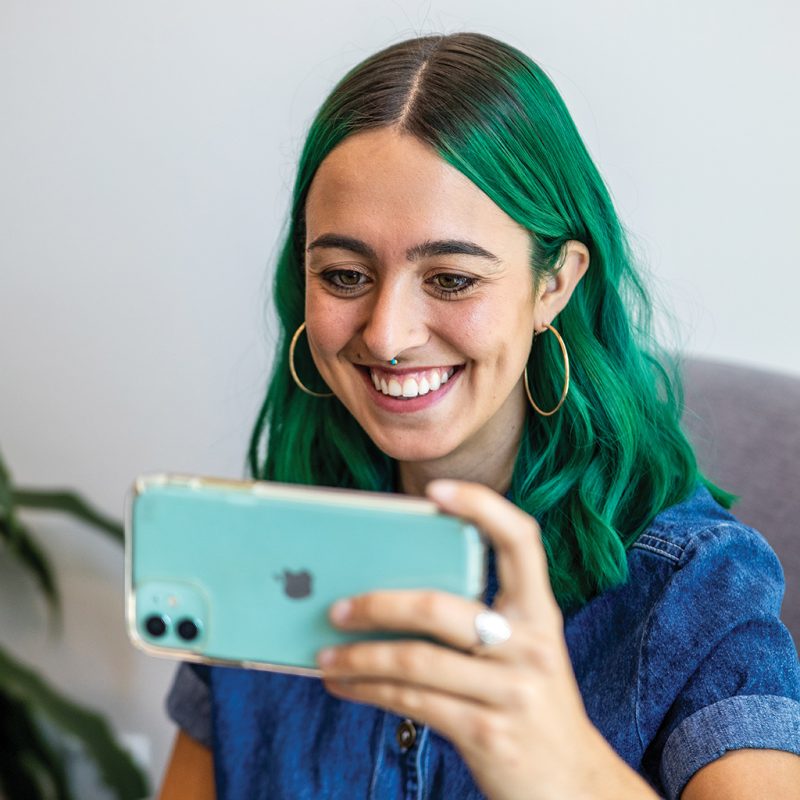 A person with long blue-green hair smiles at her phone during a telehealth appointment