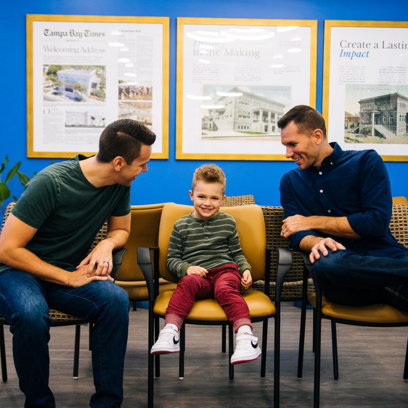 A cheerful moment captured inside a room with bright blue walls where two men and a young child are seated on chairs, engaging happily with one another. The men appear to be looking at the child with affection and pride. The walls are adorned with framed articles featuring the achievements of Metro Inclusive Health, contributing to an atmosphere of accomplishment and community support.