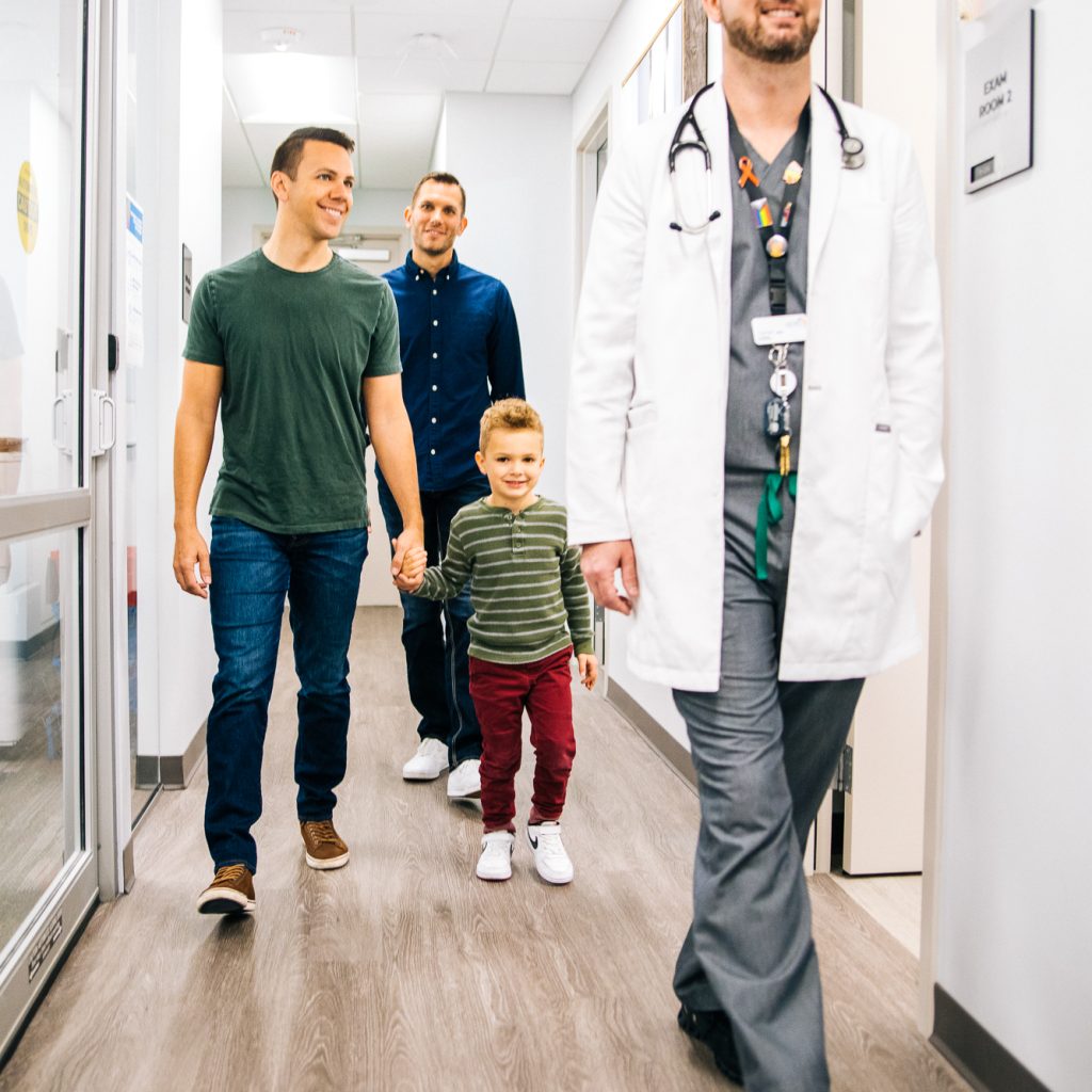In a bright healthcare facility hallway, a young boy confidently walks holding hands with two male adults, one on each side. They are followed by a healthcare professional wearing a white coat with a stethoscope and a badge adorned with pride colors. This image captures a moment of care and support, indicative of the family-friendly services at Metro Inclusive Health.