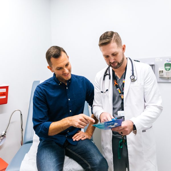 A man wearing a blue shirt learns about HIV treatment and primary care at Metro Inclusive Health. A HIV primary care provider holds information about PrEP and medical care.