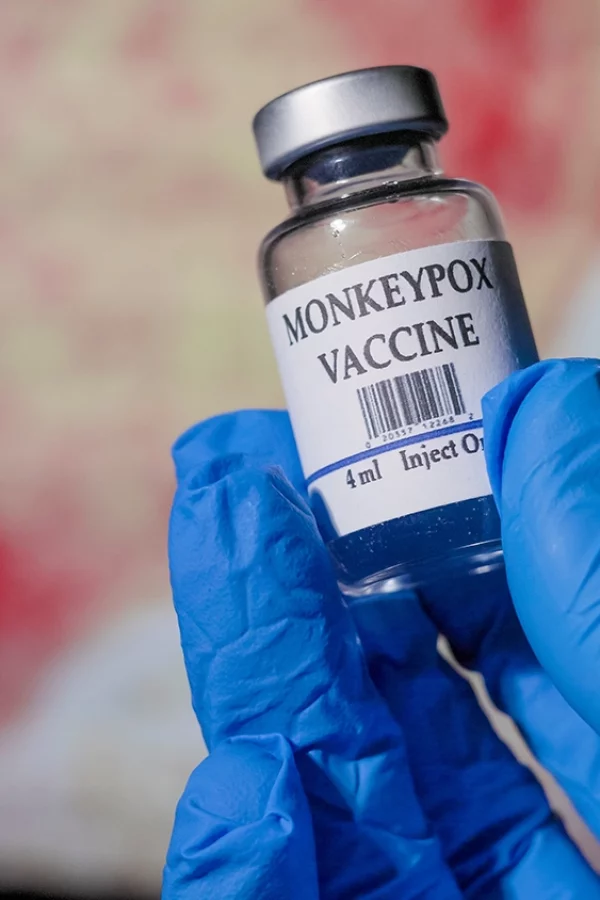 A healthcare professional wearing blue gloves holds a vial labeled 'MONKEYPOX VACCINE' in a clinical setting. The background is intentionally blurred but suggests the shape of a heart, symbolizing care and health. The vial is clear with a white label, and the text is in bold, indicating the contents for vaccination purposes.
