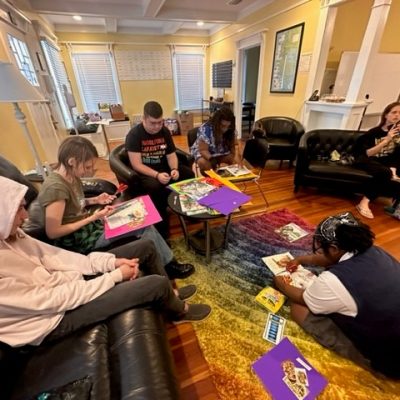A group of individuals engaged in creative activities in a cozy, well-lit room with yellow walls and wooden floors. They are seated around a coffee table, working on colorful posters, with art supplies scattered around. The room exudes a sense of community and collaboration, reflective of METRO Inclusive Health's commitment to providing welcoming spaces for activities and support.